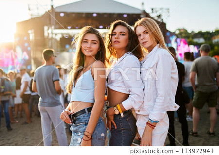 Three girls enjoy summer music festival on sandy beach at sunset. Friends pose for photo, outdoor concert stage in background. Sunlight reflects on carefree young women, crowd gathers for event. 113972941