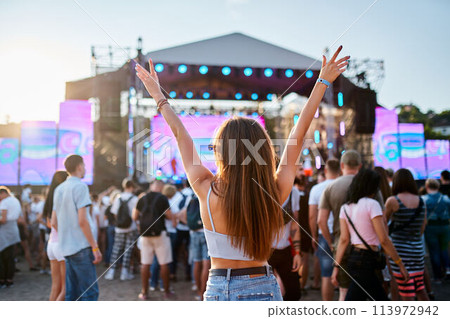 Back view of girl with raised arms, enjoying live music at sunset beach festival. Friends dance, celebrate in crowd, summer vibe at coastal event. Teens in casual wear, fun at sunny seaside gig. 113972942