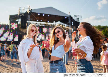 Group of girls enjoy summer music festival on sandy beach. Smiling female friends with sunglasses hold cold drinks, dance near stage in sunny day. Party, fun, youth lifestyle at sea shore event. Group of girls enjoy summer music festival on sandy beach. Smiling female friends with sunglasses hold cold drinks, dance near stage in sunny day. Party, fun, youth lifestyle at sea shore event. 113972945