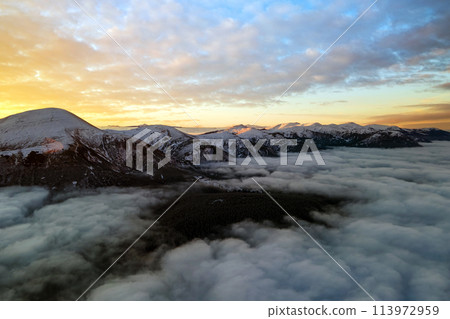 Aerial view of vibrant sunrise over white dense clouds with distant dark mountains on horizon. 113972959
