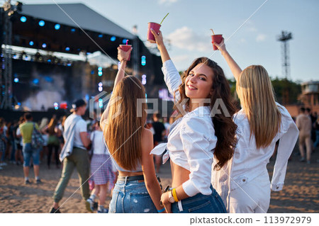 Girls with drinks enjoy live music at sunny beach festival. Group of friends dance, celebrate with cocktails. Summer party vibe on sandy shore. Attendees immerse in concert atmosphere. Girls with drinks enjoy live music at sunny beach festival. Group of friends dance, celebrate with cocktails. Summer party vibe on sandy shore. Attendees immerse in concert atmosphere. 113972979