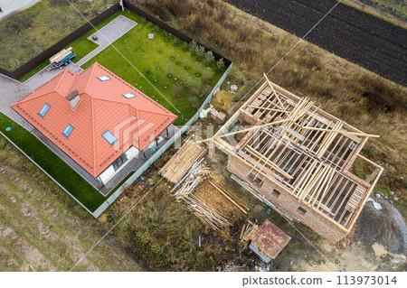 Top down aerial view of two private houses, one under construction with wooden roofing frame and another finished with red tiled roof. Top down aerial view of two private houses, one under construction with wooden roofing frame and another finished with red tiled roof. 113973014