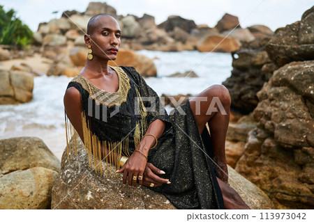 Non-binary black person in luxury dress on rocks in ocean. Trans ethnic fashion model wearing jewellery dressed in posh gown poses gracefully in tropical seaside location portrait. Pride month. 113973042