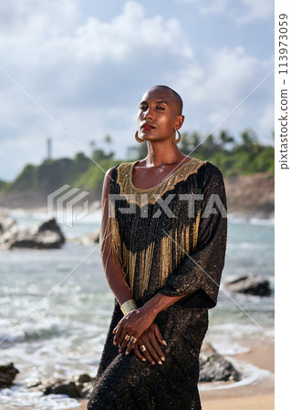 Outrageous gay black person in luxury gown poses on scenic ocean beach. Non-binary ethnic fashion model in posh outfit with makeup stands gracefully at picturesque location with sea, lighthouse. Outrageous gay black person in luxury gown poses on scenic ocean beach. Non-binary ethnic fashion model in posh outfit with makeup stands gracefully at picturesque location with sea, lighthouse. 113973059
