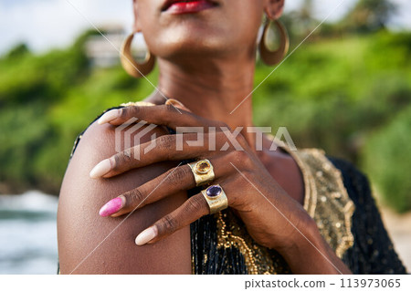 Trans sexual ethnic fashion model with brass jewelry accessories in elegant posture. Epatage gay black man with golden rings, earrings, bracelets touches shoulder on scenic ocean beach. Trans sexual ethnic fashion model with brass jewelry accessories in elegant posture. Epatage gay black man with golden rings, earrings, bracelets touches shoulder on scenic ocean beach. 113973065