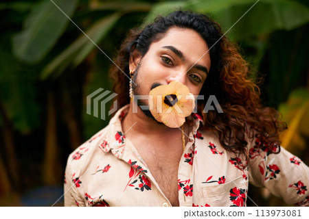 South Asian man shirt, expressive eyes holds yellow flower in mouth, bold femininity, self-love in rich garden. Person represents LGBTQ pride, cultural diversity, gender fluidity vibrant foliage. 113973081