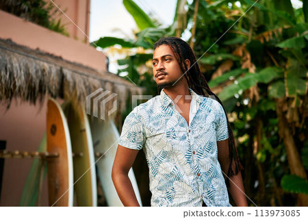 Dark-skinned surfing coach standing by thatched hut, surfboards lined up. Calm, confident instructor in tropical leaf print shirt awaits surf camp tourists. Sunset beach session forthcoming, waves. Dark-skinned surfing coach standing by thatched hut, surfboards lined up. Calm, confident instructor in tropical leaf print shirt awaits surf camp tourists. Sunset beach session forthcoming, waves. 113973085