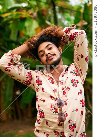 Confident gay man of color poses with flair in floral shirt amidst plants. Hands rest on head, gaze direct, stance exudes pride, freedom. Outdoor natural setting enhances subject vibrant look. Confident gay man of color poses with flair in floral shirt amidst plants. Hands rest on head, gaze direct, stance exudes pride, freedom. Outdoor natural setting enhances subject vibrant look. 113973086