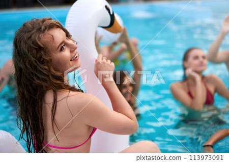 Pretty woman in bikini swimsuit hanging out on inflatable white swan mattress on night pool party. Friends partying with cocktails in holiday villa. 113973091