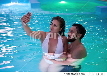 Girl in bikini hanging out, flirting with boyfriend on night pool party. Couple kisses having fun at night. Lovers doing selfie photo in swimwear on honeymoon tropical vacation. Family on holidays. 113973100