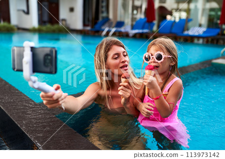 Mother and daughter enjoy ice cream in pool, selfie stick in hand, capturing smiles beside blue water. Mom and girl in swimwear share sweet moment, taking summer memories with smartphone. 113973142
