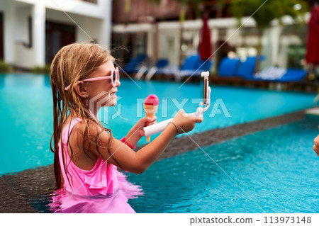 Smiling girl in pink swimsuit takes selfie in pool, holding ice cream. Child enjoys summer day, captures moment with smartphone on selfie stick. Bright, sunny leisure activity, kid at resort. Smiling girl in pink swimsuit takes selfie in pool, holding ice cream. Child enjoys summer day, captures moment with smartphone on selfie stick. Bright, sunny leisure activity, kid at resort. 113973148