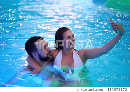 Girl in bikini hanging out, flirting with boyfriend on night pool party. Couple kisses having fun at night. Lovers doing selfie photo in swimwear on honeymoon tropical vacation. Family on holidays. 113973170