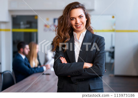 Confident businesswoman stands in office, arms crossed, smiling at camera. Colleagues in background hold a meeting. Professional, leadership, teamwork. Female executive, modern work environment. Confident businesswoman stands in office, arms crossed, smiling at camera. Colleagues in background hold a meeting. Professional, leadership, teamwork. Female executive, modern work environment. 113973186