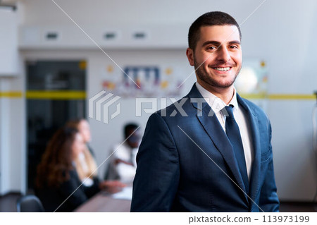 Confident eastern businessman smiles in modern office, diverse team works behind. Arab professional in formal suit leads multicultural group. Inclusive work environment, leadership, and cooperation. 113973199