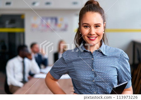Confident asian businesswoman in smart casual attire, tablet in hand, stands in modern office. Team collaboration in background with male, female coworkers engaging in discussion. 113973226