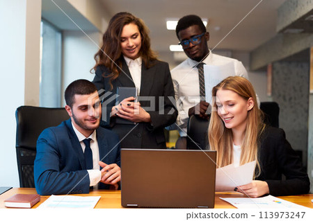 Multiracial business team collaborates around laptop in office. Smiling woman types, African observe data, suggesting successful venture. Positive pro focus on project, global diversity, teamwork. 113973247