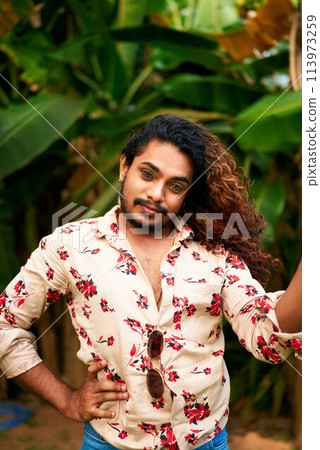 Confident gay man with wavy hair poses in tropical garden, vibrant floral shirt open, stylish sunglasses hang from collar. Hand on hip, looks directly, proud LGBTQ person with fierce expression. 113973259
