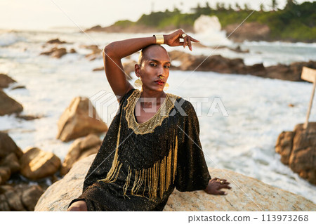Non-binary ethnic fashion model in dress, brass jewelry sits on rocks by ocean. Trans sexual black person with rings, nose-ring, bracelets, earrings in posh clothes poses in tropical seaside location. Non-binary ethnic fashion model in dress, brass jewelry sits on rocks by ocean. Trans sexual black person with rings, nose-ring, bracelets, earrings in posh clothes poses in tropical seaside location. 113973268