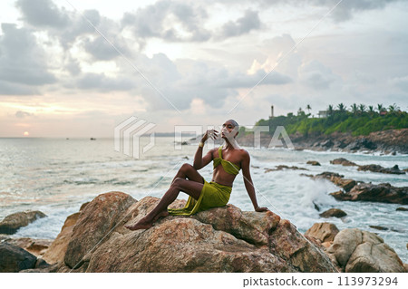 Gender fluid black person poses gracefully sitting on rocks in ocean at sunset. Androgynous slim ethnic fashion model in tropical maxi dress on top of rocky hill above storm at dusk. Pride LGBTQIA 113973294