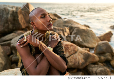 Queer black person in luxury dress, jewelry sits on rocks in ocean. Lgbtq ethnic fashion model wearing jewellery dressed in posh gown poses gracefully in tropical seaside location. Pride LGBTQIA Queer black person in luxury dress, jewelry sits on rocks in ocean. Lgbtq ethnic fashion model wearing jewellery dressed in posh gown poses gracefully in tropical seaside location. Pride LGBTQIA 113973301