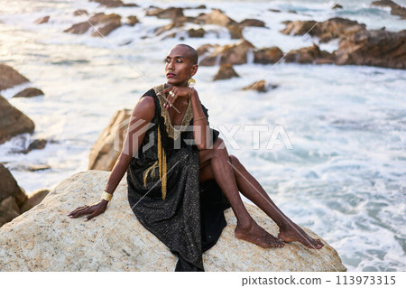 Non-binary ethnic fashion model in dress, brass jewelry sits on rocks by ocean. Trans sexual black person with rings, nose-ring, bracelets, earrings in posh clothes poses in tropical seaside location. Non-binary ethnic fashion model in dress, brass jewelry sits on rocks by ocean. Trans sexual black person with rings, nose-ring, bracelets, earrings in posh clothes poses in tropical seaside location. 113973315