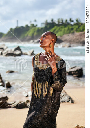Trans sexual ethnic fashion model in long posh dress and accessories in elegant posture touches neck. Epatage gay black man in luxury gown poses, touches face on scenic ocean beach. Pride month. Trans sexual ethnic fashion model in long posh dress and accessories in elegant posture touches neck. Epatage gay black man in luxury gown poses, touches face on scenic ocean beach. Pride month. 113973324