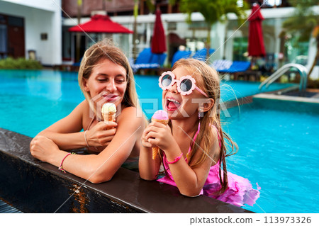 Happy girl in pool eats vanilla ice cream beside smiling mom. Summer day, child with goggles enjoys dessert, family vacation. Mother, daughter cool off, relax in water with sweet treats. 113973326