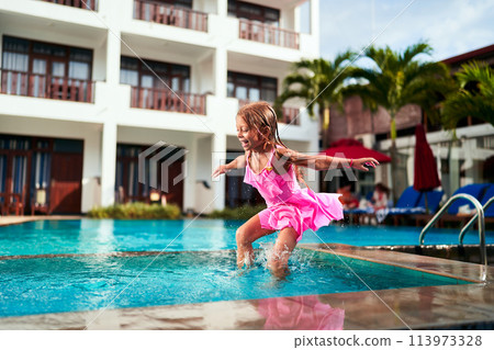 Happy girl in pink dress enjoys summer fun, jumping into outdoor pool at tropical resort. Child plays, splashes water in sunlight. Active kid vacation, leisure activity, swimming, joy, childhood. Happy girl in pink dress enjoys summer fun, jumping into outdoor pool at tropical resort. Child plays, splashes water in sunlight. Active kid vacation, leisure activity, swimming, joy, childhood. 113973328