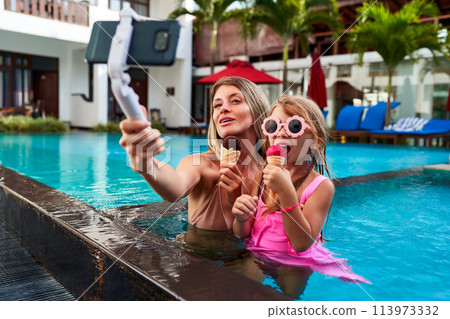 Mother and daughter enjoy summer day in pool with selfie stick, capturing moment while eating ice cream. Woman in swimsuit with girl in pink, smiles at camera, leisure fun, family vacation bonding. 113973332