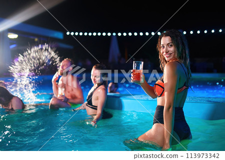 Pretty woman in bikini drinking cocktail and hanging out with friends at night pool party. People having fun dancing with drinks and splashing water in luxury private villa swimming pool. 113973342