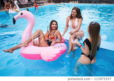 Pretty woman in bikini swimwear hanging out on inflatable pink flamingo mattress on night pool party. Friends partying with cocktails in resort villa swimming pool. Girls toast, cheer with drinks. 113973357