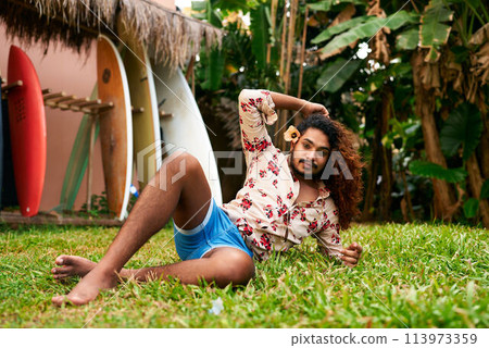 Vibrant gay man with curly hair poses in tropical garden, colorful surfboards in background. He wears floral shirt, blue shorts, outdoor fashion. LGBT traveler enjoys exotic destination, relaxed vibe. 113973359