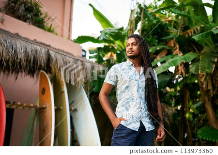 Confident dark-skinned surf instructor stands among colorful boards in tropical setting. BIPOC coach awaits students for lesson by lush green plants. Surf camp mentor ready for day training sessions. 113973360