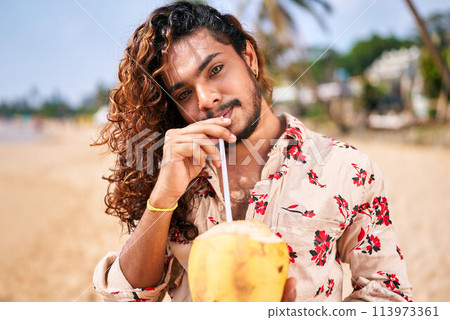 Gay man enjoys fresh coconut drink on sunny beach. Smiles, relaxed pose, tropical vacation vibe. LGBT travel lifestyle, solo male on exotic coast drinks healthy, natural beverage. Warm, clear day. 113973361