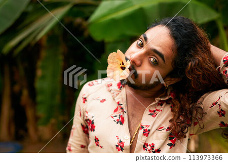 South Asian man with expressive eyes, flower in mouth, models in tropical setting. Flamboyant gay individual explores femininity, cultural identity through bold fashion, natural backdrop. South Asian man with expressive eyes, flower in mouth, models in tropical setting. Flamboyant gay individual explores femininity, cultural identity through bold fashion, natural backdrop. 113973366