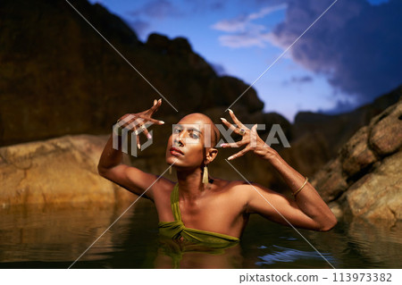 Non-binary person of color poses in water, shows jewelry inside scenic creek at night. Lgbtq ethnic graceful fashion model wears brass rings with gems, nose-ring, earrings, bracelets immersed in pond. 113973382