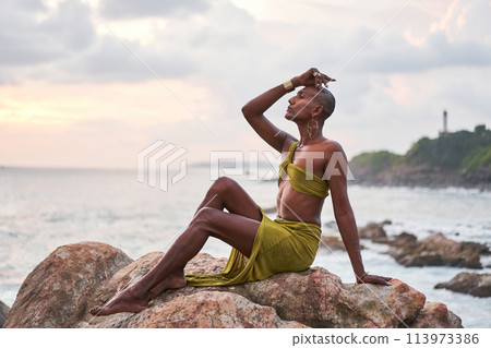 Gender fluid black person poses gracefully sitting on high rock above ocean at sunset. Androgynous ethnic fashion model in dress on rocky hill by storm at dusk. Pride month diversity concept. 113973386