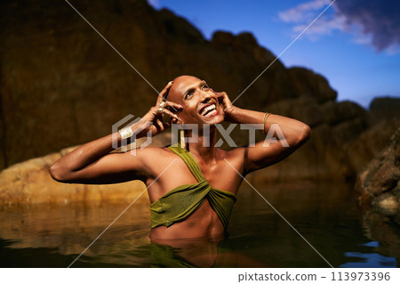 Smiling gender fluid black person poses in natural still water pool. Queer ethnic fashion model in open dress, brass jewelry with gems standing gracefully in the middle of rocky lake at night. Smiling gender fluid black person poses in natural still water pool. Queer ethnic fashion model in open dress, brass jewelry with gems standing gracefully in the middle of rocky lake at night. 113973396
