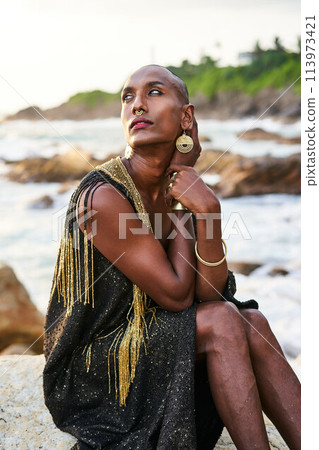 Non-binary ethnic fashion model in dress, brass jewelry sits on rocks by ocean. Trans sexual black person with rings, nose-ring, bracelets, earrings in posh clothes poses in tropical seaside location. Non-binary ethnic fashion model in dress, brass jewelry sits on rocks by ocean. Trans sexual black person with rings, nose-ring, bracelets, earrings in posh clothes poses in tropical seaside location. 113973421