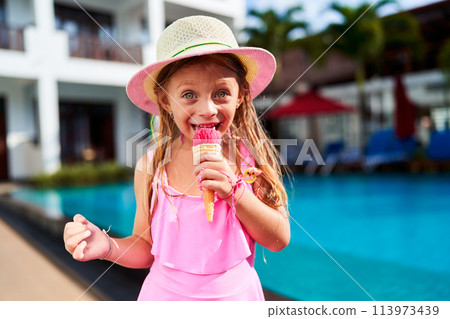 Little girl in pink swimwear and sun hat enjoys strawberry ice cream by bright blue poolside. Child with delightful expression savors summer treat, vacation ambiance, warm sunny day, leisure time. 113973439