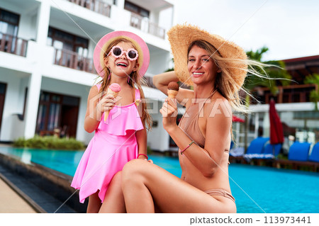 Little girl in pink dress, sunglasses enjoys ice cream by pool with smiling mom in straw hat. Summer fun, child, mother eat cold treats, relax at poolside. Happy family moment, sunshine, vacation. 113973441