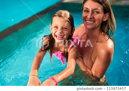 Smiling girl in pink swimsuit being taught to swim by her mom in pool. Child learns floating, kicking with mother support. Family enjoy summer swim lesson. Active lifestyle, parent-child bonding time. Smiling girl in pink swimsuit being taught to swim by her mom in pool. Child learns floating, kicking with mother support. Family enjoy summer swim lesson. Active lifestyle, parent-child bonding time. 113973457