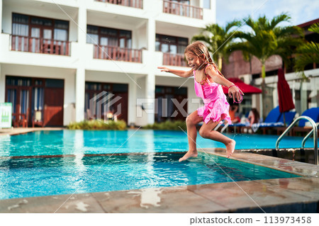 Excited young girl in pink swimsuit jumps into outdoor pool at sunny resort. Happiness, active vacation, water play depicted. Childhood, summer, leisure, travel, luxury hotel backdrop. 113973458