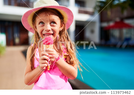 Smiling girl in pink dress, straw hat enjoys strawberry ice cream by poolside. Child relishes frozen treat on sunny day, outdoor summer setting. Innocent youth leisure, refreshing vacation activity. 113973462