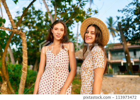 Two young women in vintage polka dot and floral dresses pose at tropical beach resort, summer vibe with retro fashion, friends smile near palm trees enjoying sunset, holiday relaxation, style concept. 113973472