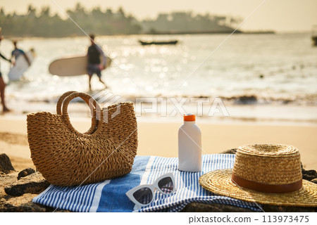 Beach scene with straw bag, sunscreen bottle, on blue towel. Sunglasses rest beside, essentials for sun safety, relaxation by sea. Sunny ocean backdrop with people, surfboard hint at active leisure. Beach scene with straw bag, sunscreen bottle, on blue towel. Sunglasses rest beside, essentials for sun safety, relaxation by sea. Sunny ocean backdrop with people, surfboard hint at active leisure. 113973475