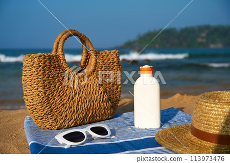 Woven tote bag stands beside sunscreen bottle on striped blue towel at sunny tropical beach. Sun hat and white sunglasses rest on sand, essential gear for safe sunbathing near ocean waves. Woven tote bag stands beside sunscreen bottle on striped blue towel at sunny tropical beach. Sun hat and white sunglasses rest on sand, essential gear for safe sunbathing near ocean waves. 113973476
