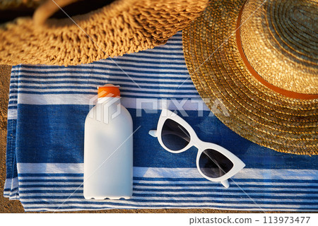 Overhead shot of summer essentials on sandy shore, white sunscreen bottle next to chic sunglasses, beside striped towel under woven sunhat for stylish sun protection at beach 113973477