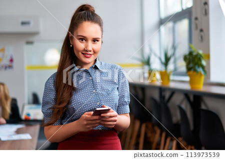 Asian businesswoman in smart casual attire uses smartphone in modern office, colleagues in background. Pro conducts fintech operations, engages in corporate communication, manages digital. 113973539